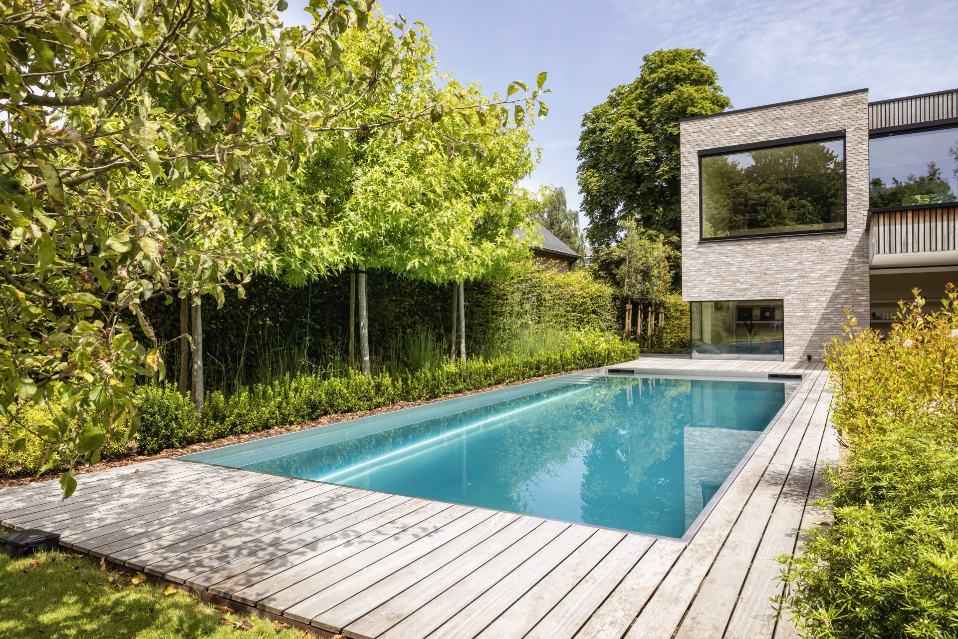 piscina in acciaio inox 10x4 m: vista laterale della piscina in acciaio inox con siepi verdi, prato e facciata in mattoni, Luxury, Design, Exclusive, Modern, Custom Made, Special, Beautiful