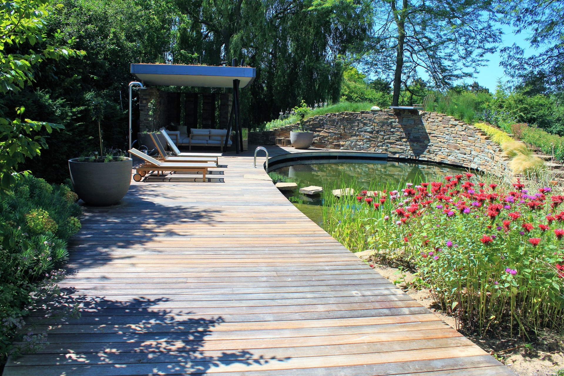vasca da nuoto naturale con cascata: vista lungo la terrazza in legno accanto alla vasca da nuoto con fioritura e muro in, Luxury, Design, Exclusive, Modern, Custom Made, Special, Beautiful